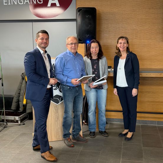 Vier Menschen stehen zusammen und lächeln für ein Foto. Sie halten Papiere und ein Buch. Hinter ihnen befindet sich eine Holzwand mit einem Schild, das 'Eingang A' besagt. In der Nähe steht ein Lautsprecher und ein Stativ.