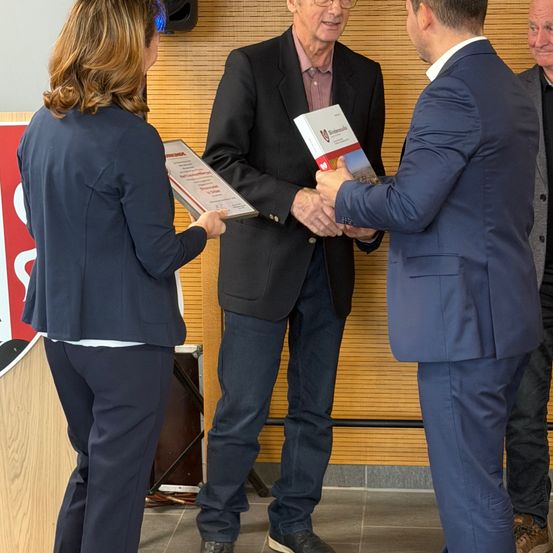 Ein Mann, der ein Buch in der Hand hält, gibt einem anderen Mann die Hand. Eine Frau steht neben ihm und hält ein Papier. Ein weiterer Mann steht hinter ihnen und schaut zu.
