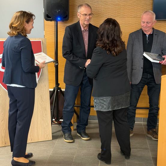 Vier Personen, zwei Männer und zwei Frauen, stehen zusammen in einem Raum und halten Urkunden. Eine Frau hält eine Urkunde in der linken Hand und schaut die andere Frau an. Ein Mann in der Mitte schüttelt einer Frau die Hand. Ein Lautsprecher ist auf einem Stativ montiert.