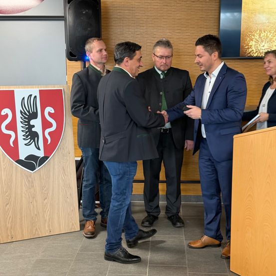 Eine Gruppe formell gekleideter Männer in einem Raum schütteln sich in einem Raum die Hände vor einem hölzernen Podium mit einem Logo. Dahinter befindet sich eine hölzerne Wand und ein Fernseher.