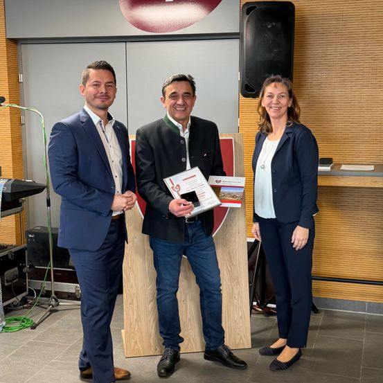 Drei Personen stehen zusammen und lächeln für ein Foto. Ein Mann hält ein Zertifikat, während zwei andere zusehen und neben einem Podium mit einem Schild stehen. Ein Lautsprecher befindet sich an der Wand hinter ihnen.