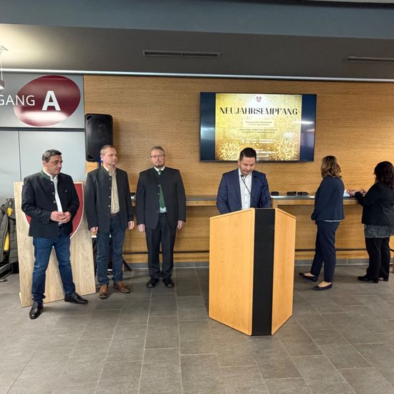 Fünf Männer und zwei Frauen stehen hinter einem Podium, an dem ein Mann in ein Mikrofon spricht. Hinter ihm befindet sich eine große Leinwand, auf der 'NEUJAHRESEMPFANG' steht.