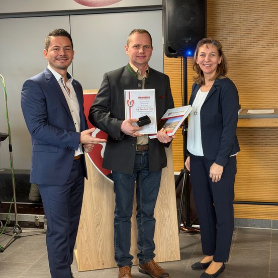 Drei Menschen stehen lächelnd vor einem hölzernen Podium. Der Mann in der Mitte hält ein Zertifikat und ein Buch. Die Frau auf der rechten Seite hält ein Buch. Dahinter steht ein Lautsprecher.
