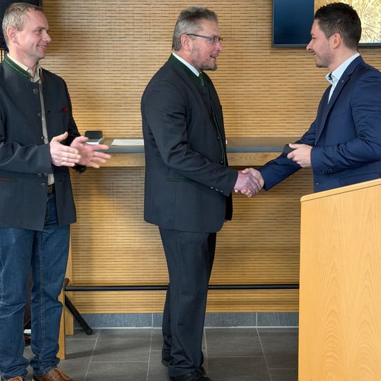 Drei Männer stehen in einem Innenraum, zwei von ihnen schütteln sich die Hände. Ein Podest steht vor ihnen. Sie lächeln und scheinen miteinander zu sprechen. Ein Monitor ist an der Wand hinter ihnen montiert.