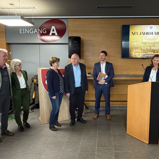 Mehrere Personen stehen in der Nähe eines Podiums, einer Mann hält ein Buch und ein anderer steht an einem Mikrofon. Hinter ihnen steht ein großes Schild mit der Aufschrift 'EINGANG A', und ein Bildschirm zeigt 'NEUJAHRSSEM'. Die Umgebung wirkt feierlich.