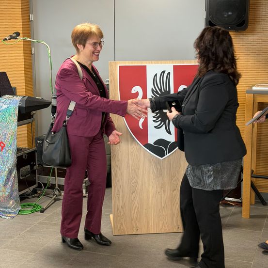 Zwei Frauen schütteln sich die Hände vor einem hölzernen Podium mit einem großen roten und weißen Wappen darauf.