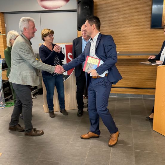 Zwei Männer schütteln sich die Hände in einem Raum, während andere zuschauen. Ein Mann hält ein Buch. Die Umgebung ist mit Holzwänden ausgestattet.