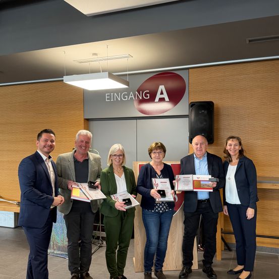Fünf Personen stehen nebeneinander, alle lächeln und halten Auszeichnungen. Sie befinden sich in einem Raum mit Holzwänden und einem Schild, das 'EINGANG A' besagt. Hinter ihnen steht ein Lautsprecher und ein Holztisch.