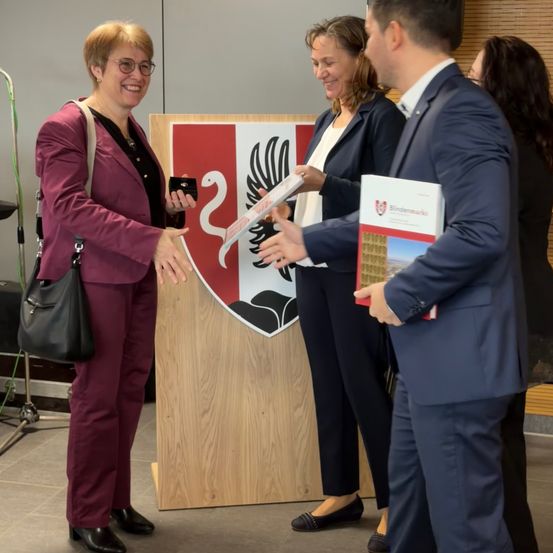Drei Personen stehen vor einem hölzernen Podium mit einem rot-weißen Wappen. Die Frau auf der linken Seite lächelt, während sie ein kleines Objekt hält, und der Mann auf der rechten Seite hält ein Buch. Die Frau in der Mitte lächelt und schaut auf die Frau auf der linken Seite.