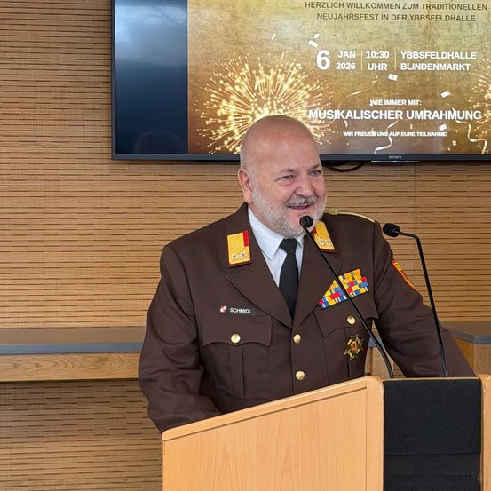 Ein Militärbeamter steht lächelnd am Rednerpult mit einem Mikrofon. Hinter ihm zeigt eine Leinwand ein Feuerwerk und Text für eine Neujahrsveranstaltung.