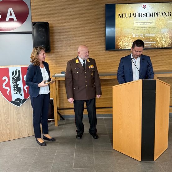 Drei Personen stehen in einem Raum mit einem Podium. Ein Mann hält eine Rede am Podium.