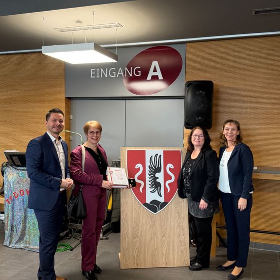 Fünf Personen stehen vor einem Podest mit einem Schild, eine Frau überreicht einer anderen Frau ein Zertifikat. Dahinter befindet sich eine Holzwand mit einem Schild, auf dem 'Eingang A' steht.