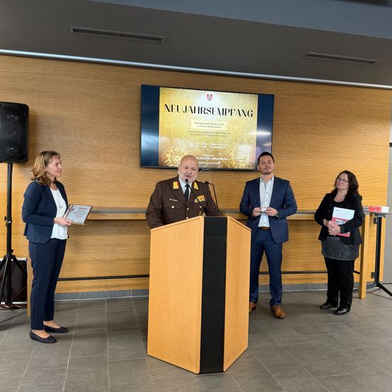Ein Militäroffizier hält eine Rede an einem Podium vor einem Monitor. Zwei Frauen und ein Mann stehen neben ihm. Eine Frau hält ein Zertifikat.
