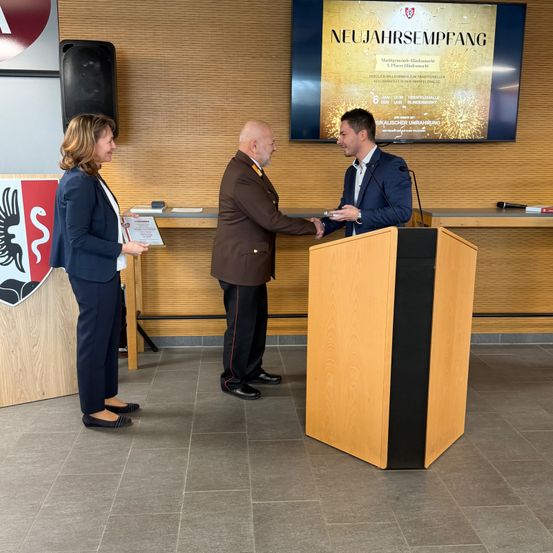 Eine Frau und zwei Männer stehen vor einem Podium. Der Mann auf der rechten Seite schüttelt dem Mann in der Mitte die Hand. Hinter ihnen zeigt ein Monitor 'NEUAJHARSEMPFANG' an.