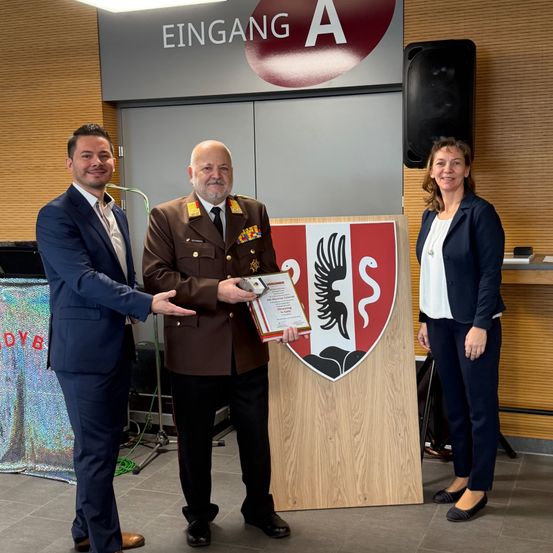 Drei Personen stehen vor einem Holzschild mit einem rot-weißen Wappen, möglicherweise bei einer Preisverleihung. Ein Mann in Uniform hält ein Zertifikat, während die anderen lächeln. Dahinter ist ein Lautsprecher an der Wand montiert.