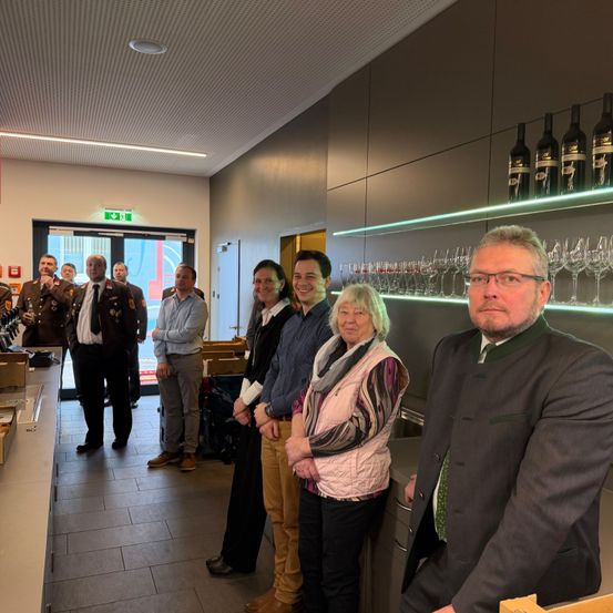 Eine Gruppe von Menschen, darunter Personen in Uniformen, steht in einem Raum mit weißer Decke, gefliestem Boden und einer Wand mit Regalen, die Weinflaschen und Weingläser halten.