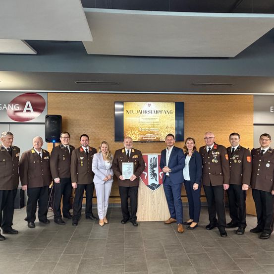 Eine Gruppe von Militärangehörigen steht in einem Innenraum. Eine Frau und ein Mann in der Mitte halten eine Plakette. Hinter ihnen zeigt ein Monitor ein goldenes Poster. Vor ihnen steht ein hölzerner Podest mit einem Schild.