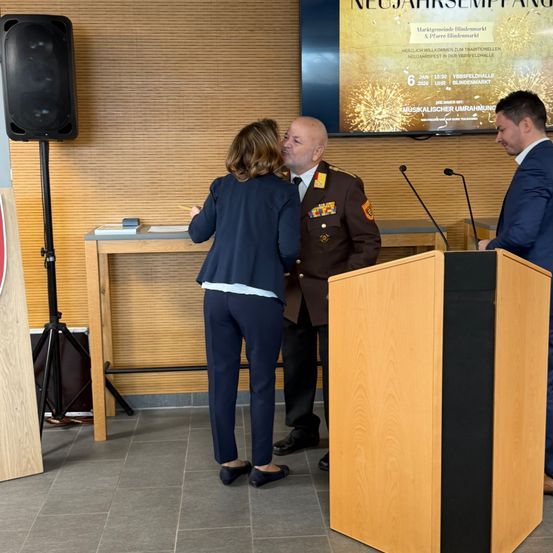 Eine Frau in einem blauen Anzug küsst einen Mann in Uniform, während ein anderer Mann an einem Rednerpult steht. Hinter ihnen zeigt ein großer Monitor eine Silvesterfeier.