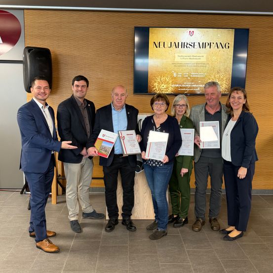 Eine Gruppe von sechs Personen in formeller Kleidung steht zusammen und posiert möglicherweise für ein Foto. Sie halten Urkunden in den Händen und lächeln. Die Umgebung scheint ein formelles Ereignis zu sein. An der Wand hinter ihnen befindet sich ein großes Schild mit der Aufschrift 'NEUJAHRSEMPPFANG'.