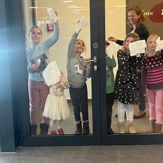 Eine Gruppe junger Mädchen und eine Frau stehen hinter einer Glastür und halten Taschentücher in der Hand, möglicherweise feiern sie einen besonderen Anlass.