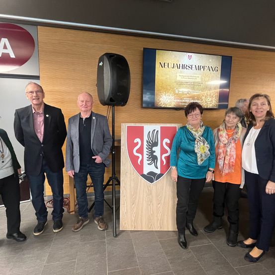 Mehrere Menschen stehen zusammen in einem Raum und posieren für ein Foto. Vor ihnen steht ein hölzerner Podest mit einem Wappen.