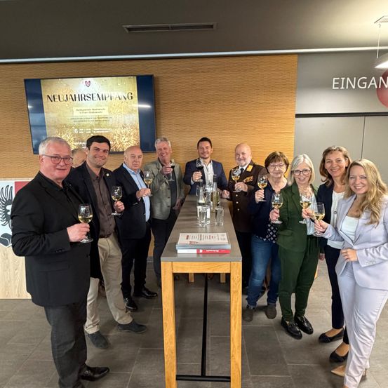Eine Gruppe von Menschen in formeller Kleidung steht um einen langen Tisch herum und hält Weingläser in der Hand. Im Hintergrund befindet sich eine Wand mit einem großen Schild, das 'EINGAN' und einem kleineren Schild, das 'NEUJAHRSEMPSANG' steht.