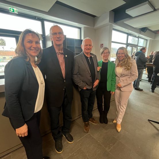 Fünf Personen stehen in einem Raum und lächeln für ein Foto. Sie tragen formelle Kleidung. Hinter ihnen sind große Fenster und Menschen, die stehen und reden.