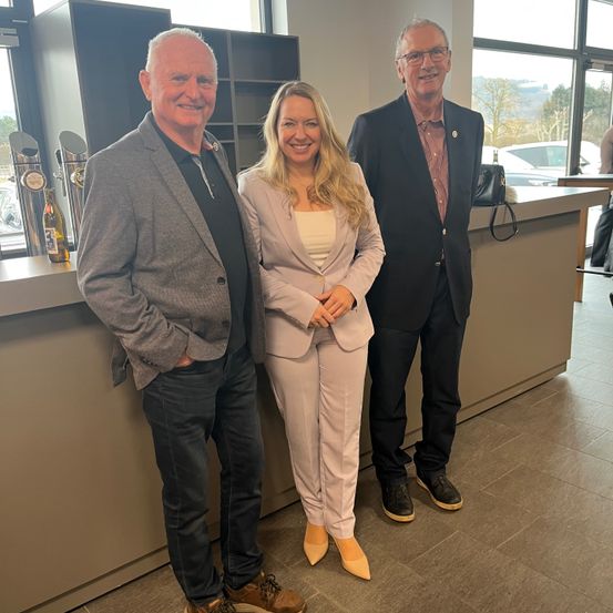 Drei Menschen stehen vor einer Theke, zwei Männer und eine Frau, lächeln in die Kamera. Dahinter sind Regale und ein Fenster zu sehen.