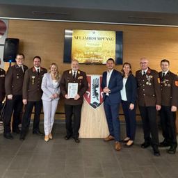 Eine Gruppe uniformierter Personen, sowohl Männer als auch Frauen, stehen zusammen vor einer Wand mit einem Schild, das 'NEUJAHRSEEMPFANG' besagt. Ein Mann hält ein Zertifikat. Dahinter ist ein Lautsprecher an der Wand montiert.