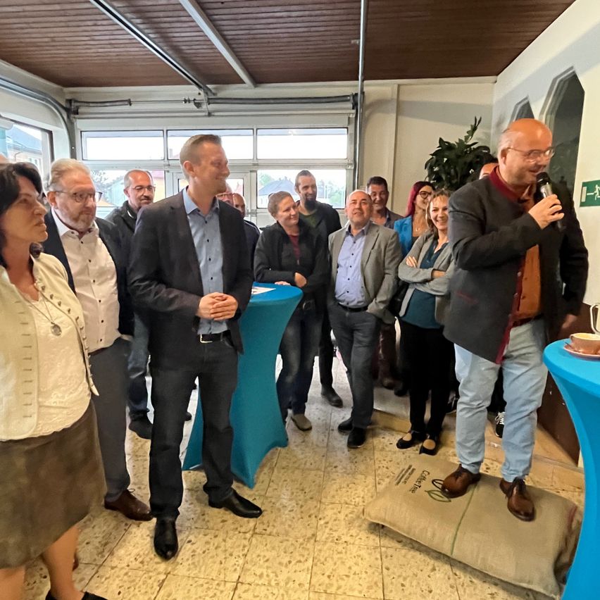 Eine Gruppe von Menschen in formeller Kleidung steht in einem Raum mit Tischen, ein Mann auf einem Beanbag spricht in ein Mikrofon