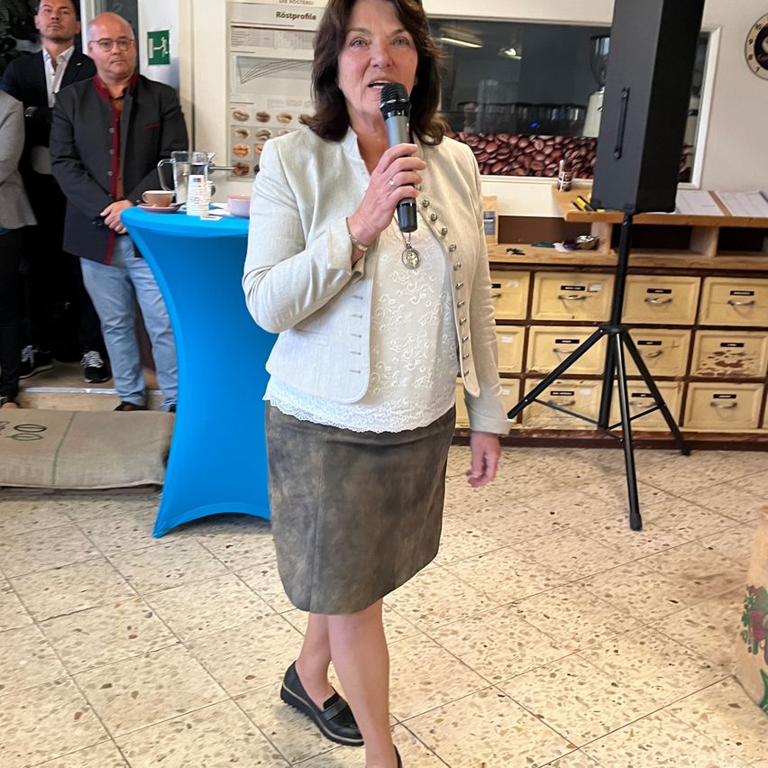 Eine Frau mit einem Mikrofon steht vor einem blauen Tisch und mehreren Personen. Sie trägt eine weiße Jacke und einen grauen Rock.