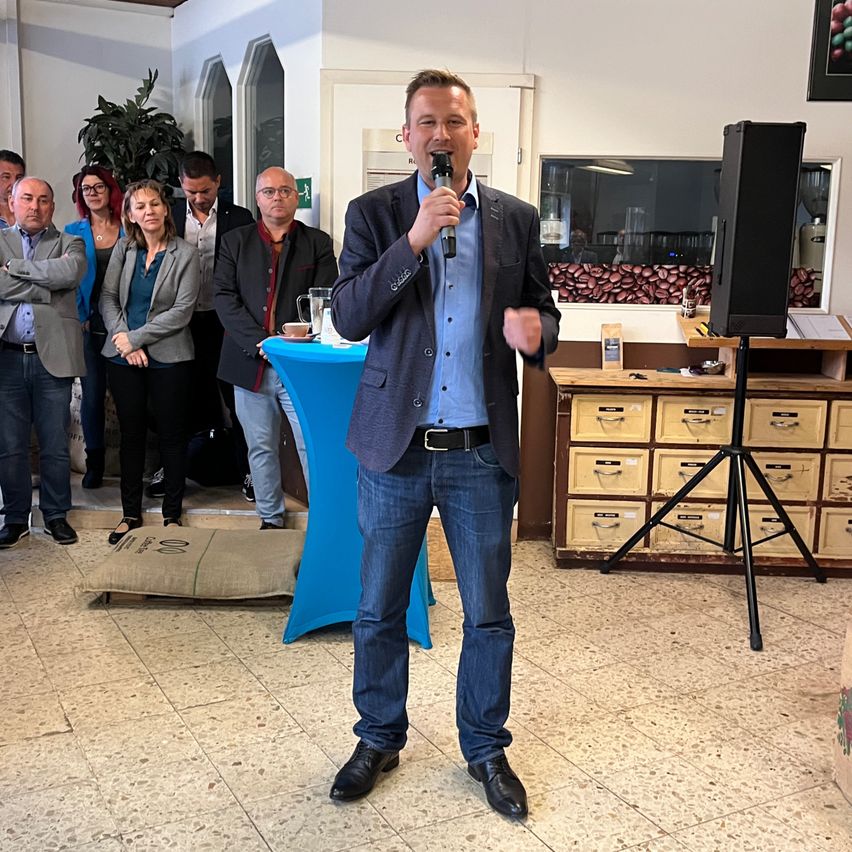 Ein Mann spricht in ein Mikrofon bei einer Versammlung, mit einem Tisch und Holzschubladen im Hintergrund.