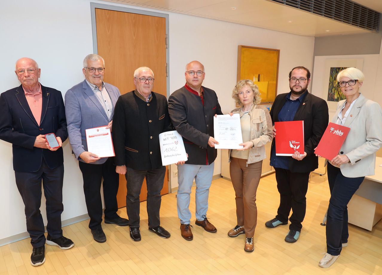 Eine Gruppe von Personen, die Urkunden und Dokumente halten, möglicherweise bei einer Zeremonie. Sie stehen in einem Raum mit Holzboden.