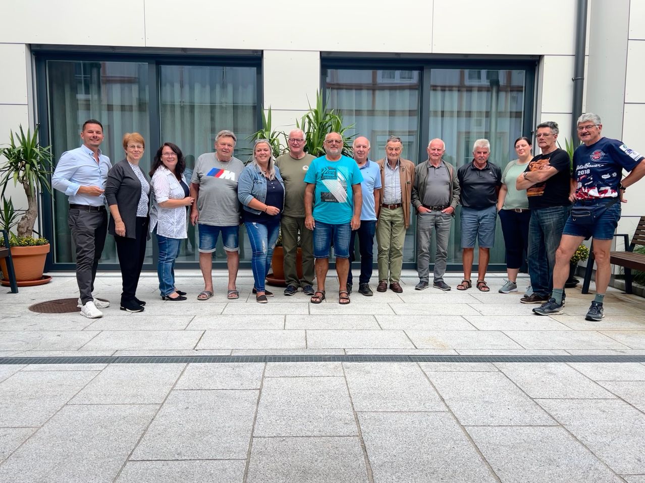 Eine Gruppe von Menschen in lässiger Kleidung steht vor einem modernen Gebäude mit Glasfenstern und einem Topf mit Pflanzen. Sie scheinen für ein Foto zu posieren.