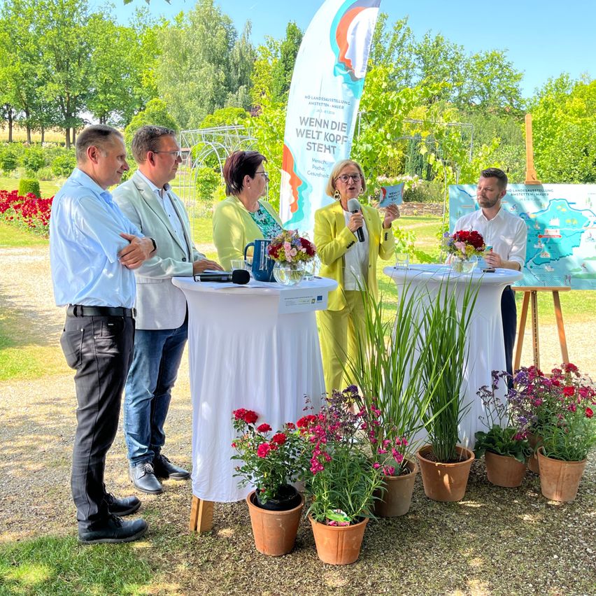 Fünf Personen stehen neben Tischen mit Blumen, eine hält ein Mikrofon. Sie befinden sich wahrscheinlich bei einer Gartenveranstaltung. Dahinter steht ein großes Banner und eine Karte.