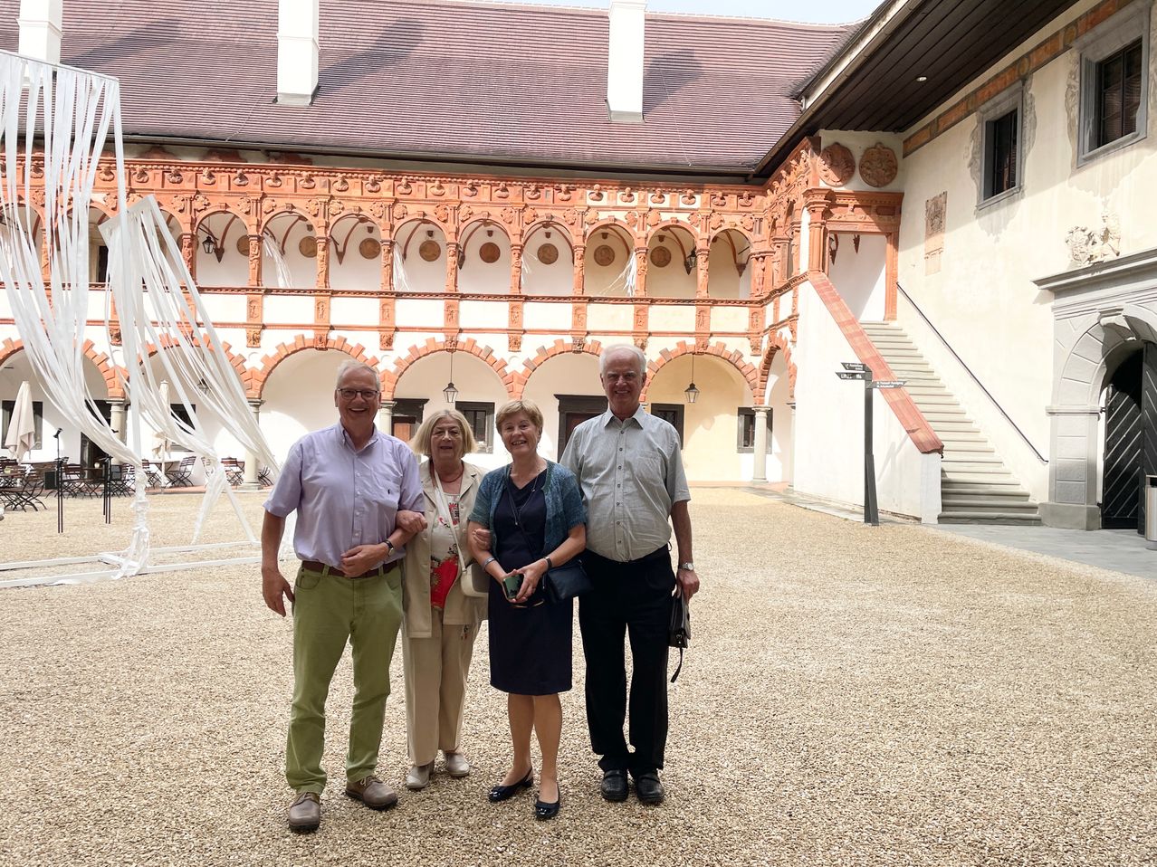 Vier Erwachsene stehen lächelnd vor einem Gebäude mit verzierten Bögen und machen ein Foto. Der Mann auf der linken Seite trägt eine Brille und einen Gürtel. Die Frau in der Mitte hält ein kleines Objekt in der Hand. Die Frau auf der rechten Seite trägt eine Halskette. Das Gebäude hat eine Treppe auf der rechten Seite.