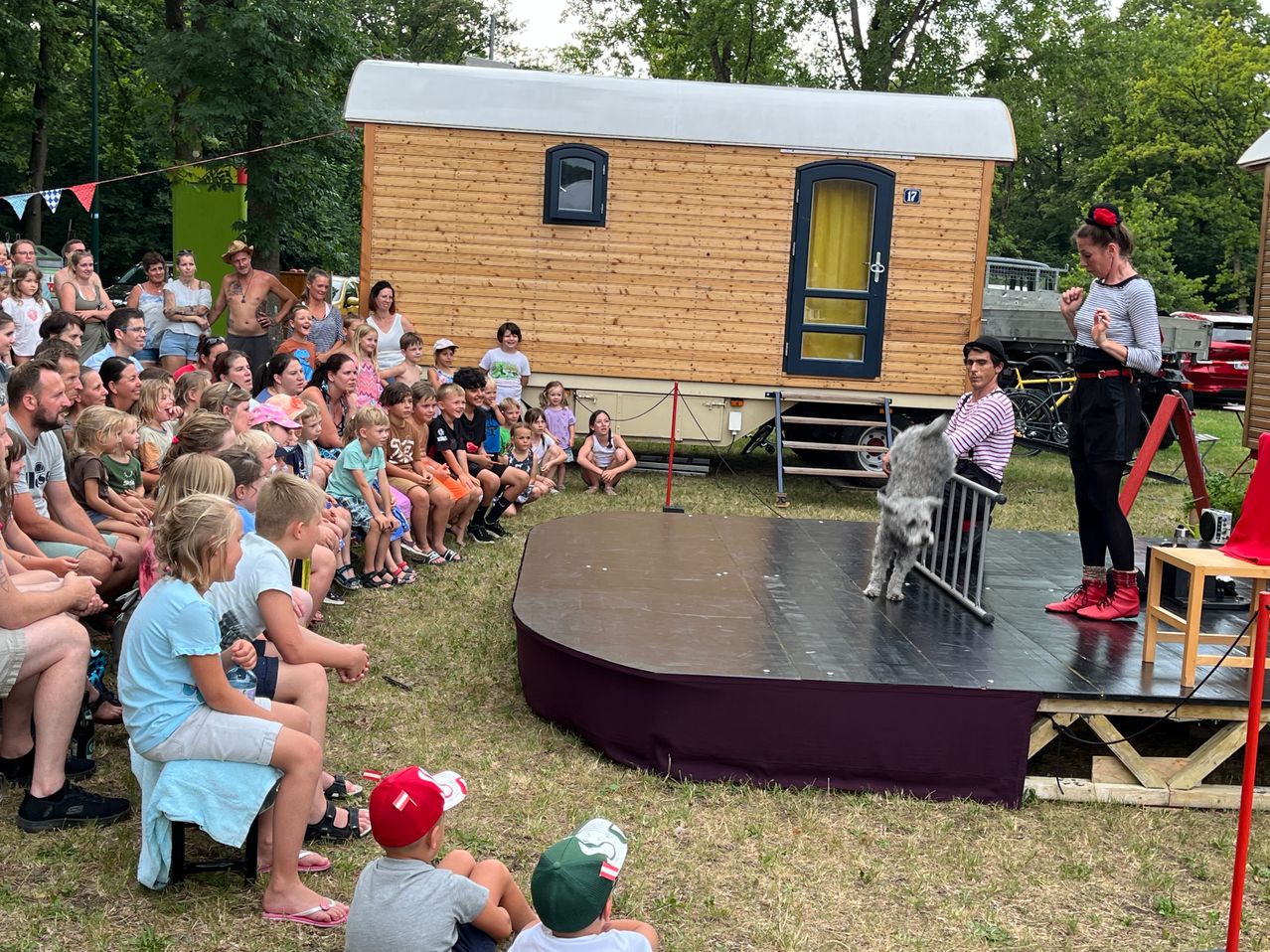 Eine Gruppe von Kindern und Erwachsenen sitzt um eine Bühne herum und beobachtet, wie ein Hund Tricks vorführt. Die Bühne hat einen schwarzen und lila Rand. Dahinter steht ein Wohnwagen mit Fenster und Tür auf Gras.