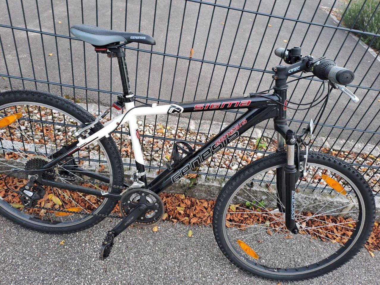 Ein schwarz-weißes Mountainbike steht neben einem Metallzaun. Es hat das Wort Sigma auf dem Rahmen, mit Genesis auf dem Sattel und den Pedalen.