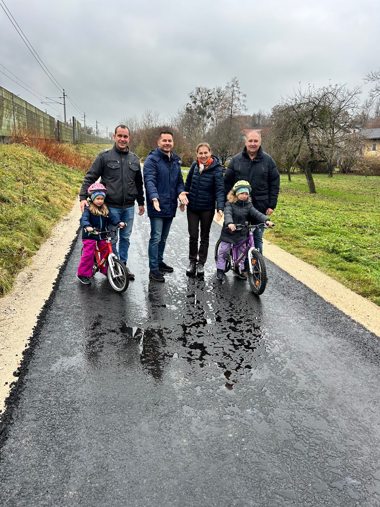 Eine Familie mit vier Personen und zwei Kindern steht auf einer Straße. Zwei Kinder fahren Fahrräder, eines rot und eines lila. Bäume und ein Zaun sind im Hintergrund.