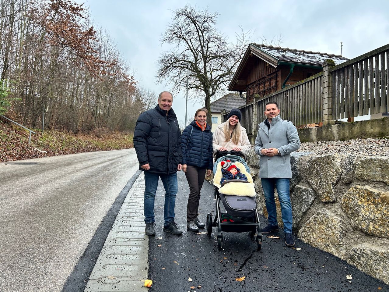 Vier Personen stehen auf einem Weg neben einer Straße. Einer von ihnen schiebt einen Kinderwagen mit einem Baby. Sie sind warm gekleidet, was auf kaltes Wetter hindeutet. Hinter ihnen steht ein Haus mit einem Zaun und einem Baum.
