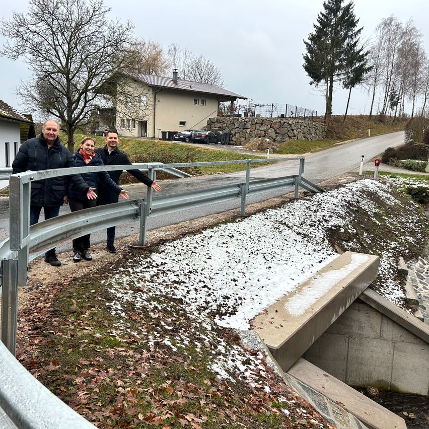 Drei Personen stehen auf einem Metallgeländer über einem schneebedeckten Bereich mit einer Betonbrücke, in der Nähe eines Hauses und einer Straße.