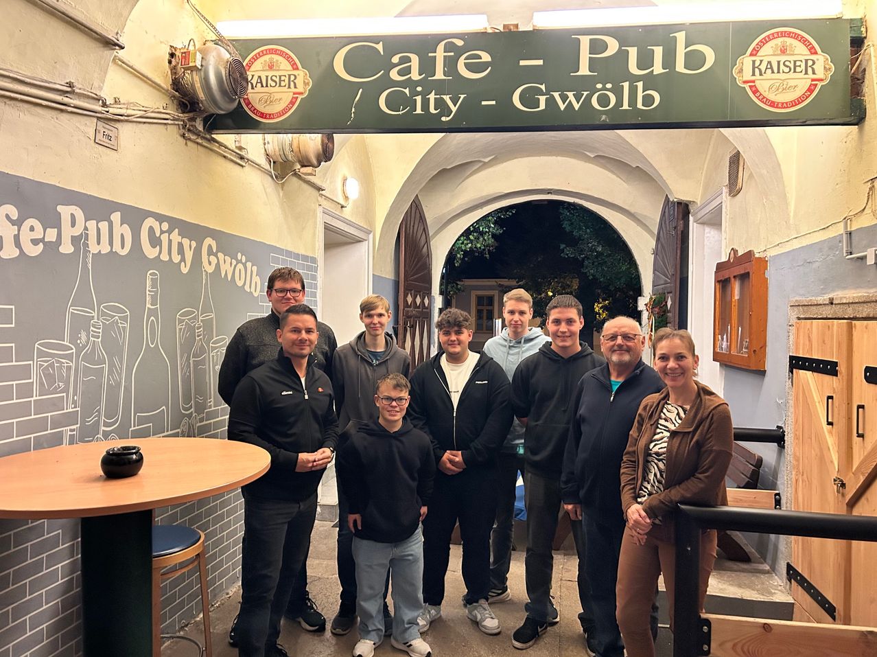 Eine Gruppe von Menschen posiert für ein Foto im Cafe-Pub City-Gwolb, mit einem Schild über ihnen. Sie tragen alle Jacken, und einige von ihnen tragen Brillen. Es gibt einen Holztisch mit einer Schüssel darauf. Die Wand hat ein Wandbild mit Flaschendesign.