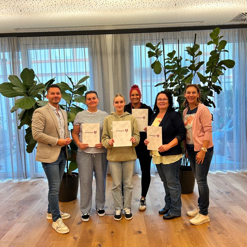 Eine Gruppe von Menschen in einem Raum mit Holzböden, die Urkunden halten. Sie stehen neben großen Topfpflanzen und weißen Vorhängen.