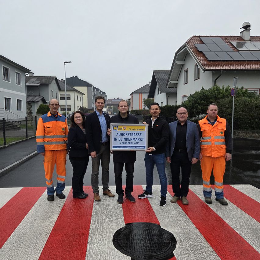 Eine Gruppe von Menschen steht auf einem Zebrastreifen und hält ein Schild mit der Aufschrift Aufhofstrasse in Blindenmarkt. Sie tragen alle orange Westen und scheinen Beamte zu sein.