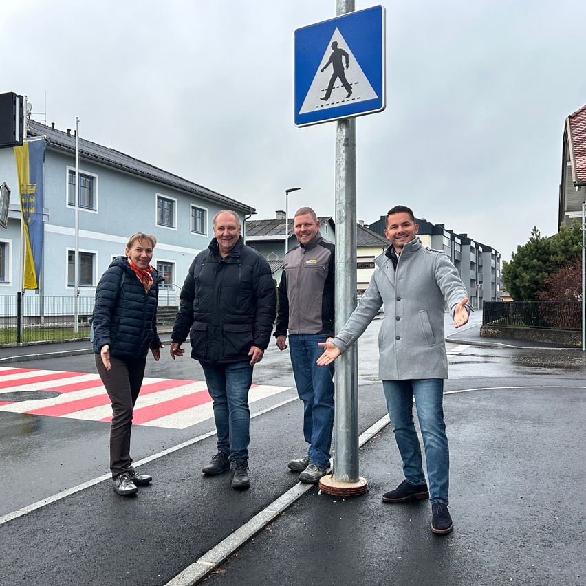 Vier Personen stehen auf einer nassen Straße mit einem Fußgängerübergangsschild vor ihnen. Dahinter befindet sich ein Gebäude mit einem Fahnenmast.