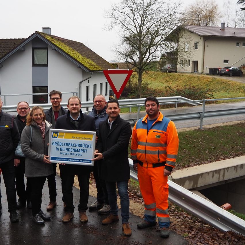 Eine Gruppe von Menschen steht draußen und hält eine große Plakette, auf der 'Groblerbachbrucke in Blindenmarkt im Zuge der L 6046' steht. Dahinter befinden sich Häuser und eine Straße.