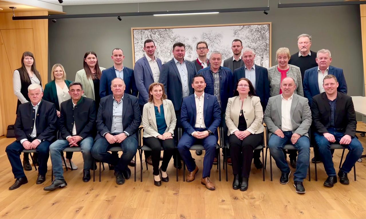 Eine Gruppe von Menschen sitzt in einem Raum. Sie tragen formelle Kleidung. Einige stehen. Sie posieren wahrscheinlich für ein Gruppenfoto. Der Raum hat Holzböden und eine Wand mit einem großen Bilderrahmen.