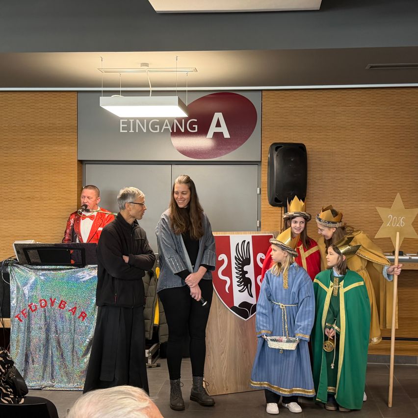 Eine Gruppe von Menschen in Kostümen steht vor einem Podium mit einem Schild, das 'Eingang A' besagt. Eine Person hält einen sternbesetzten Stab. Dahinter steht ein Mann mit Mikrofon an einem Tisch. Ein Lautsprecher ist an der Wand montiert.