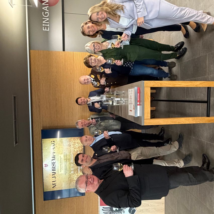 Eine Gruppe von Menschen mit Weinglas in der Hand steht vor einer Wand mit den Worten 'EINGAN'. Sie befinden sich in einem Raum mit einem Holztisch und gefliestem Boden. An der Wand hängt ein Banner mit 'NEULAHRSEMPFANG'.
