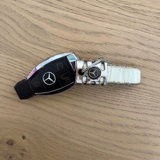 Ein Mercedes-Benz Autoschlüssel liegt auf einer Holzoberfläche. Er hat eine silberfarbene Metalloberfläche, ein schwarzes Gummipolster und einen Schlüsselanhänger.